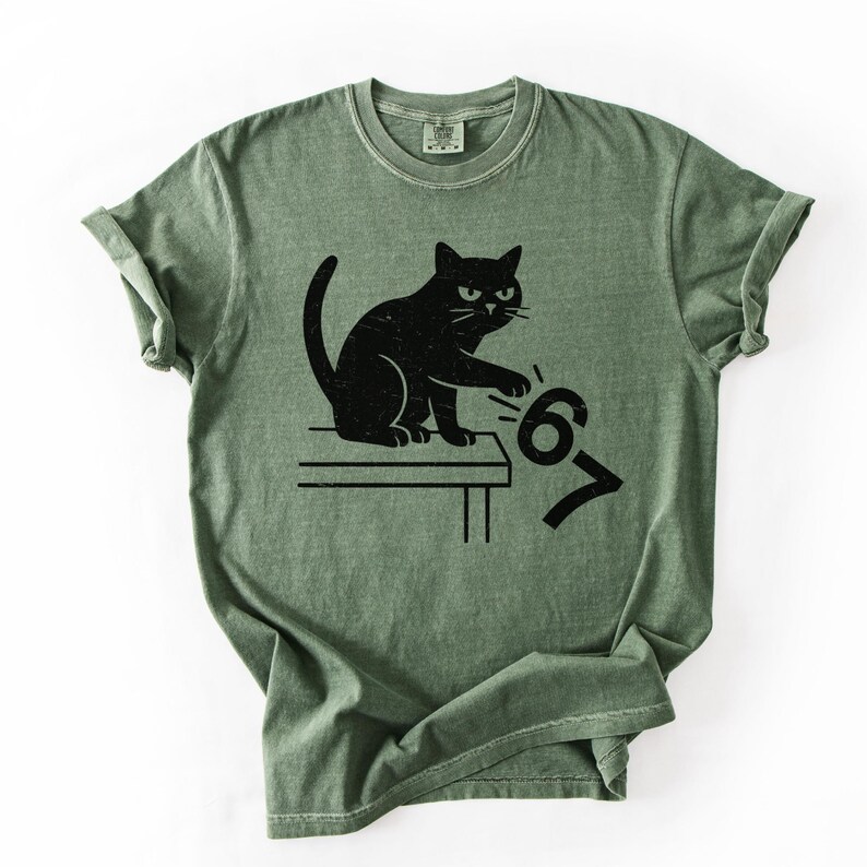 May include: Green t-shirt featuring a black cat illustration. The cat is sitting on a table and appears to be pushing the numbers "67" off the table. The shirt has a classic crew neck and short sleeves.