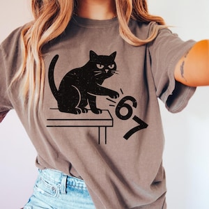 May include: A gray t-shirt featuring a black cat illustration. The cat is sitting on a table and reaching for the numbers "67". The shirt has a relaxed fit and short sleeves.