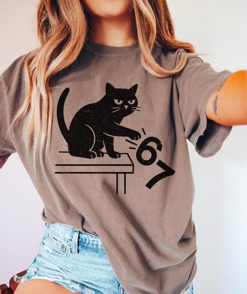 May include: A gray t-shirt featuring a black cat illustration. The cat is sitting on a table and reaching for the numbers "67". The shirt has a relaxed fit and short sleeves.