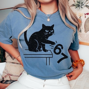 May include: A light blue t-shirt featuring a black cat graphic. The cat is sitting on a table with the numbers 6 and 7 next to it. The shirt has short sleeves and a crew neck.