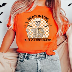 May include: Orange t-shirt with a graphic featuring a skeleton holding a coffee cup and giving a peace sign. The text reads "DEAD INSIDE BUT CAFFEINATED" in a retro font. The design includes bats and a checkered background.