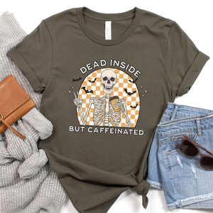 May include: Olive green t-shirt featuring a skeleton graphic holding a coffee cup, with the text "DEAD INSIDE BUT CAFFEINATED". The design includes bats and a checkered background, perfect for Halloween or a coffee lover.