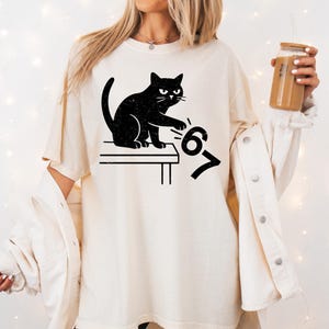 May include: Cream-colored t-shirt featuring a black cat illustration. The cat is sitting on a table, paw extended towards the numbers "6" and "7". The design has a distressed, vintage look.