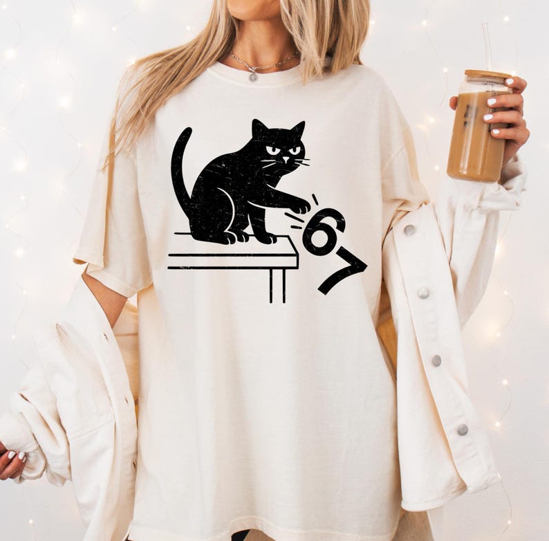 May include: Cream-colored t-shirt featuring a black cat illustration. The cat is sitting on a table, paw extended towards the numbers "6" and "7". The design has a distressed, vintage look.