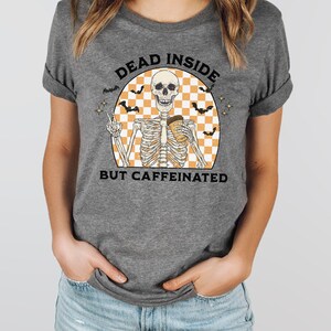 May include: Gray t-shirt featuring a skeleton graphic holding a coffee cup and making a peace sign. The graphic includes the text "DEAD INSIDE BUT CAFFEINATED" with bats and a checkered background. A fun, casual tee.