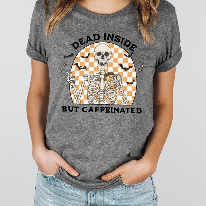 May include: Gray t-shirt featuring a skeleton graphic holding a coffee cup and making a peace sign. The graphic includes the text "DEAD INSIDE BUT CAFFEINATED" with bats and a checkered background. A fun, casual tee.