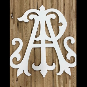 May include: White wooden monogram with the letters "A" and "S" intertwined. The letters are cut out in a decorative, ornate style. The monogram is displayed on a woodgrain background.