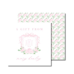 Peut inclure: Une étiquette cadeau avec un motif floral rose. L'étiquette présente le texte "A GIFT FROM" au-dessus d'un écusson rose avec les initiales "MHV" et le nom "mary hanley" en dessous. L'étiquette est décorée de fleurs roses et de feuilles vertes.