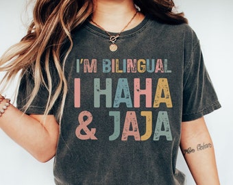 Puede incluir: Una camiseta gris oscuro con el texto "I'm Bilingual I HAHA & JAJA" impreso en letras coloridas y desgastadas.