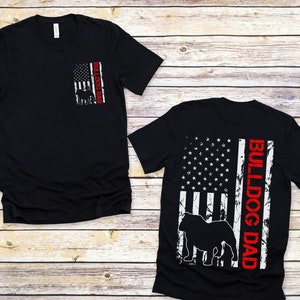 May include: Two black t-shirts featuring a distressed American flag design. One shirt has the text "Bulldog Dad" on the back, and the other has the text "Bulldog Dad" on the front pocket.