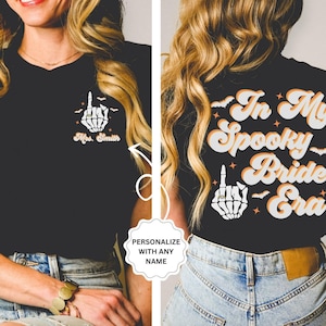 Puede incluir: Camiseta negra con una mano de esqueleto haciendo el dedo medio y el texto "In My Spooky Bride Era" en una fuente retro. La mano del esqueleto lleva un anillo. El texto "Mrs. Smith" est&aacute; en la parte delantera de la camiseta.