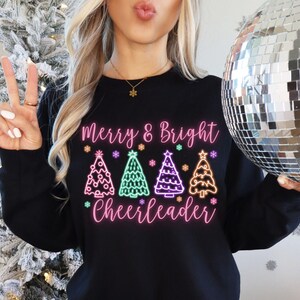 Pu&ograve; includere: Felpa nera con un design rosa e verde con quattro alberi di Natale e il testo "Merry & Bright Cheerleader".