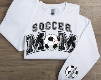 May include: White sweatshirt with black text that reads "Soccer Mom" with a black and white soccer ball with a red heart in the center. The left sleeve has a black and white soccer ball with the word "Alexa" written on it.
