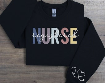 Può includere: Felpa nera con il testo "NURSE registered" in colori arcobaleno. La manica sinistra ha un contorno bianco di uno stetoscopio e un cuore.