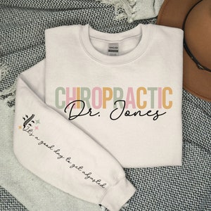 May include: A white sweatshirt with the text "CHIROPRACTIC Dr. Jones" in colorful letters. The left sleeve has a small graphic of a spine with lightning bolts and stars and the text "It's a good day to get adjusted".
