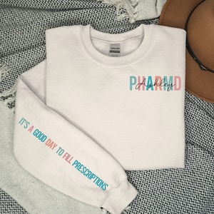 May include: White sweatshirt with the text "PHARM.D. Ashley" on the front and the text "It's a good day to fill prescriptions" on the sleeve.