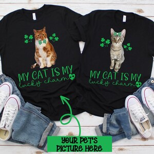 May include: Two black t-shirts with a green shamrock design and the text "My cat is my lucky charm". One shirt features a brown tabby cat wearing a bow tie, the other features a gray tabby cat wearing a green bow. The text "Your pet's picture here" is written in green on a white background.