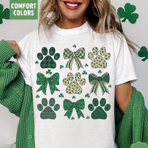 May include: White t-shirt featuring a pattern of green paw prints and bows with plaid, leopard, and solid designs. The shirt includes the text "COMFORT COLORS" and "White". The design incorporates shamrocks, suggesting a St. Patrick's Day theme.