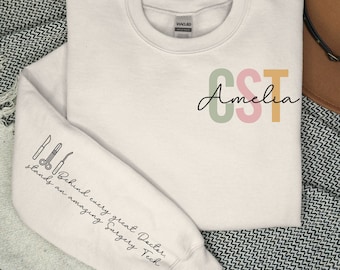 May include: A light pink sweatshirt with the text "CST Amelia" in green, pink, and yellow letters. The left sleeve has a black and white illustration of surgical tools with the text "Behind every great Doctor, stands an amazing Surgery Tech."