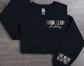 May include: Black sweatshirt with the text "BOOK CLUB Ashley" on the front and "ONE MORE CHAPTER" on the sleeve. The text is in rainbow colors.