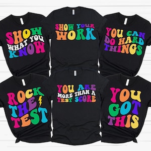 May include: Set of six black t-shirts with colorful text. The shirts feature motivational phrases like "Show Your Work", "You Are More Than A Test Score", and "You Got This".