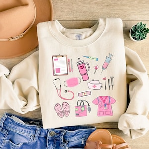 K&ouml;nnte beinhalten: Cremefarbenes Sweatshirt mit rosa medizinischen Illustrationen, darunter ein Stethoskop, eine Gesichtsmaske und medizinische Werkzeuge. Das Sweatshirt wird mit einer blauen Jeans und einem braunen Hut pr&auml;sentiert. Die Illustrationen sind im Cartoon-Stil.