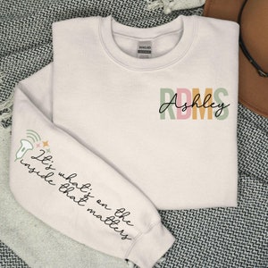 May include: A light pink sweatshirt with the text "It's what's on the inside that matters" on the left sleeve and the text "RDMS Ashley" on the right side of the chest. The text is in a colorful font.