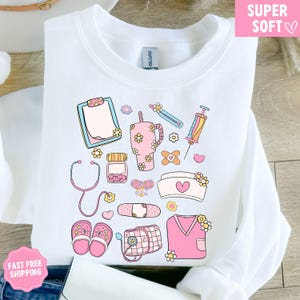 May include: White sweatshirt with a colorful medical-themed design. The design includes a clipboard, stethoscope, medicine bottle, syringe, bandages, nurse's cap, and other medical elements, all decorated with flowers. The words "SUPER SOFT" are in a pink box.