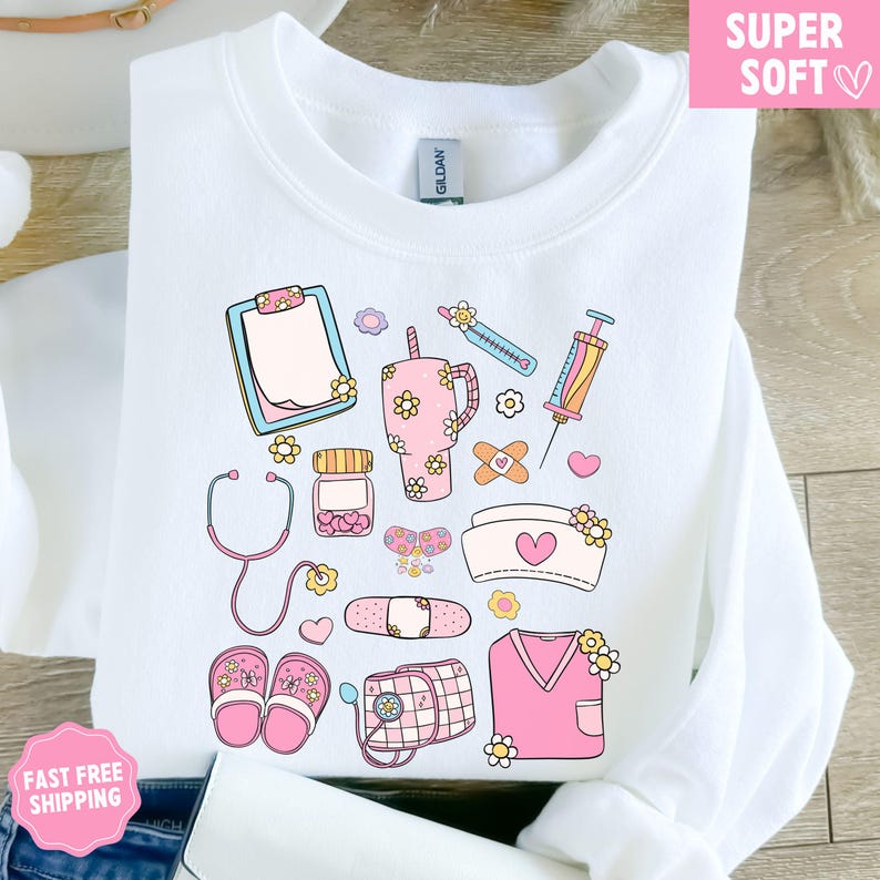 May include: White sweatshirt with a colorful medical-themed design. The design includes a clipboard, stethoscope, medicine bottle, syringe, bandages, nurse's cap, and other medical elements, all decorated with flowers. The words "SUPER SOFT" are in a pink box.