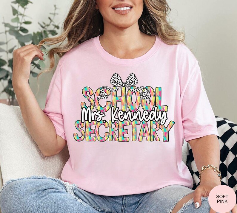 Może przedstawiać: R&oacute;żowa koszulka z tęczowym i czarno-białym wzorem w kropki z napisem "School Mrs. Kennedy Secretary".