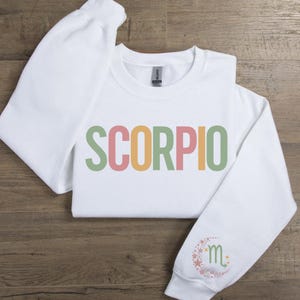 May include: White sweatshirt with the word "SCORPIO" printed in green, yellow, pink, and orange letters. The left sleeve has a small, colorful design of a crescent moon with stars and the astrological symbol for Scorpio.
