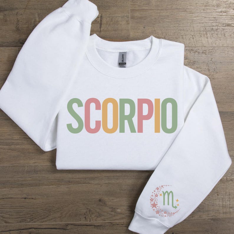 May include: White sweatshirt with the word "SCORPIO" printed in green, yellow, pink, and orange letters. The left sleeve has a small, colorful design of a crescent moon with stars and the astrological symbol for Scorpio.