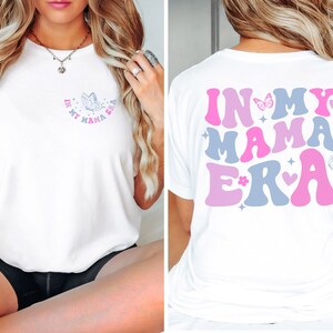 May include: White t-shirt with the text "In My Mama Era" in pink, blue, and white letters with a butterfly and hearts. The text is in a retro font.