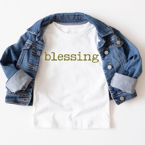 May include: White t-shirt with the word "blessing" printed in green on the front. The t-shirt is folded and laid on a white surface with a blue denim jacket draped over it.