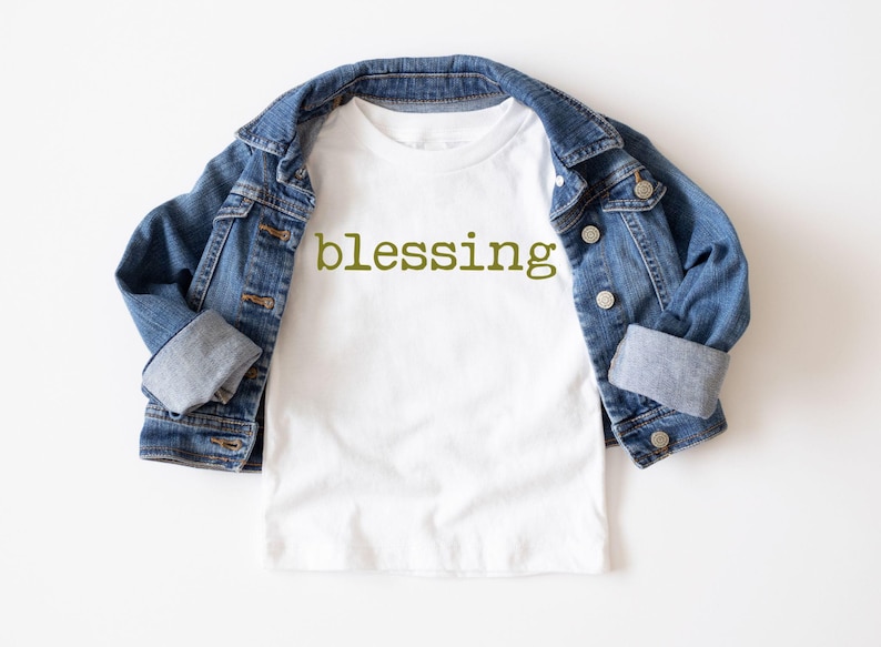 May include: White t-shirt with the word "blessing" printed in green on the front. The t-shirt is folded and laid on a white surface with a blue denim jacket draped over it.