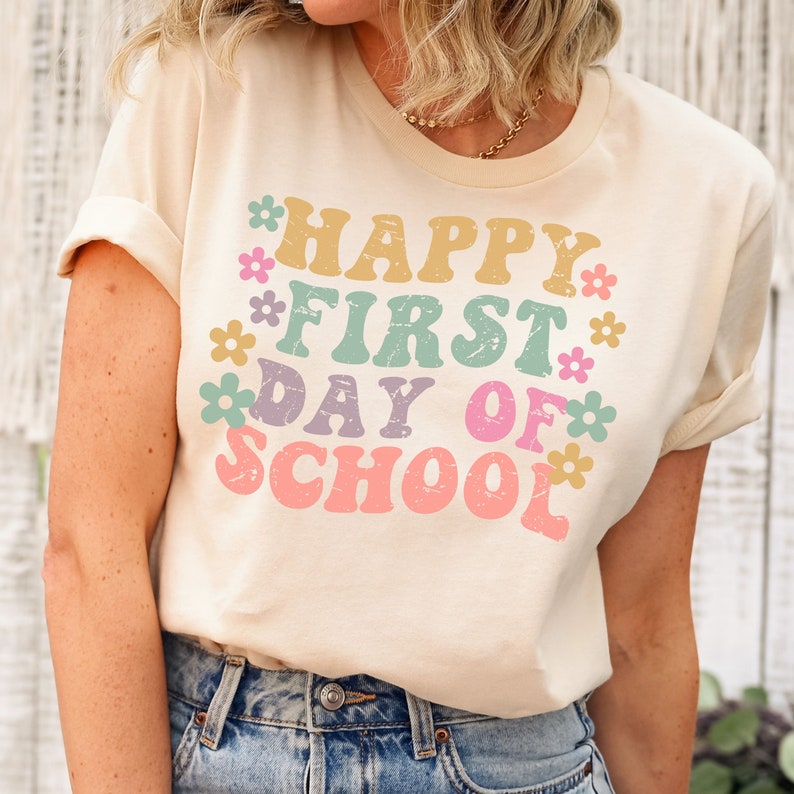 Puede incluir: Una camiseta beige claro con el texto "HAPPY FIRST DAY OF SCHOOL" en un arco&iacute;ris de colores con flores que rodean el texto.