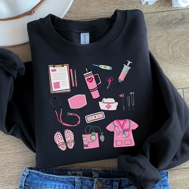 K&ouml;nnte beinhalten: Schwarzes Sweatshirt mit rosa medizinischen Illustrationen, darunter ein Stethoskop, eine Gesichtsmaske und ein Klemmbrett. Weitere Elemente sind ein Thermometer, eine Spritze, eine Krankenschwesteruniform, eine Wasserflasche, eine Schere, ein Pflaster und eine Krankenschwesterhaube. Das Sweatshirt liegt auf Denim.