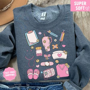 May include: A dark gray sweatshirt featuring a collection of pink and yellow medical-themed illustrations. The design includes a stethoscope, prescription bottle, bandages, a nurse's cap, and a tumbler. The words "SUPER SOFT" are in a pink box.