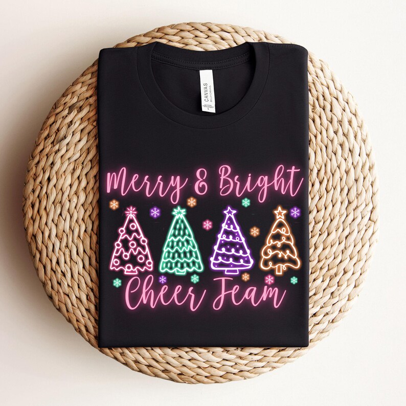 Pu&ograve; includere: Maglietta nera con un design rosa "Merry & Bright Cheer Team". Il design presenta quattro alberi di Natale colorati al neon con luci rosa, verdi, viola e arancioni.