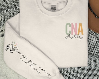 May include: A white sweatshirt with the text "CNA Ashley" in colorful letters on the front and the text "Because even nurses need heroes" on the left sleeve.