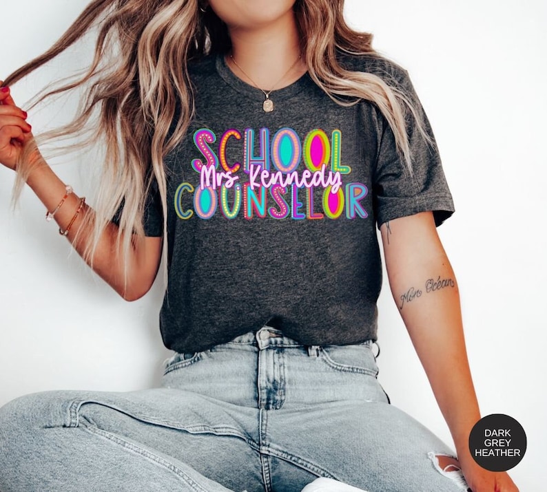 May include: A dark heather grey t-shirt with the text "SCHOOL Mrs. Kennedy COUNSELOR" in colorful, outlined letters.