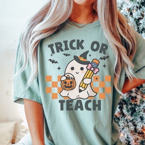 K&ouml;nnte beinhalten: Ein hellgr&uuml;nes T-Shirt mit dem Text "Trick or Teach" in Schwarz. Ein Cartoon-Geist mit einer Hexenm&uuml;tze h&auml;lt einen Bleistift und einen K&uuml;rbis-Eimer.