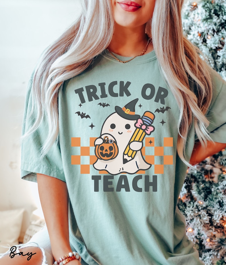 K&ouml;nnte beinhalten: Ein hellgr&uuml;nes T-Shirt mit dem Text "Trick or Teach" in Schwarz. Ein Cartoon-Geist mit einer Hexenm&uuml;tze h&auml;lt einen Bleistift und einen K&uuml;rbis-Eimer.