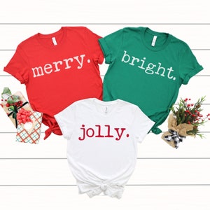 May include: Three Christmas themed t-shirts with the words "merry", "bright", and "jolly" printed on them. The shirts are red, green, and white.