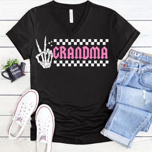 K&ouml;nnte beinhalten: Schwarzes V-Ausschnitt-T-Shirt mit einer wei&szlig;en Skeletthand, die das Peace-Zeichen zeigt, und einem rosafarbenen karierten Banner mit dem Text "Grandma".