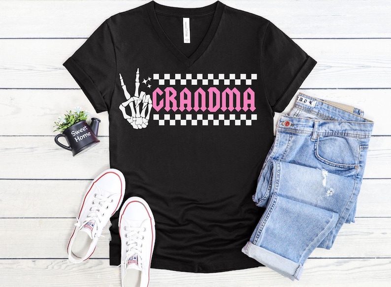 K&ouml;nnte beinhalten: Schwarzes V-Ausschnitt-T-Shirt mit einer wei&szlig;en Skeletthand, die das Peace-Zeichen zeigt, und einem rosafarbenen karierten Banner mit dem Text "Grandma".