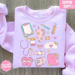 May include: A light purple sweatshirt featuring a collection of medical-themed illustrations. The design includes a stethoscope, clipboard, syringe, bandages, and a nurse's cap, all adorned with floral accents. The words "SUPER SOFT" are in a pink box.