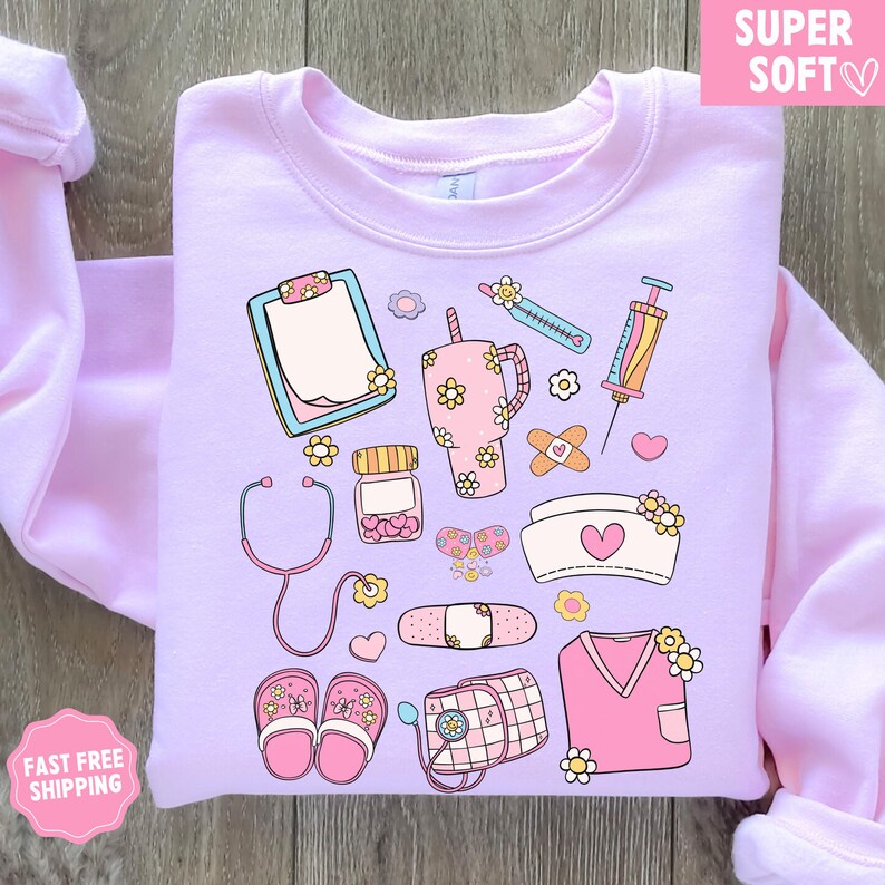 May include: A light purple sweatshirt featuring a collection of medical-themed illustrations. The design includes a stethoscope, clipboard, syringe, bandages, and a nurse's cap, all adorned with floral accents. The words "SUPER SOFT" are in a pink box.