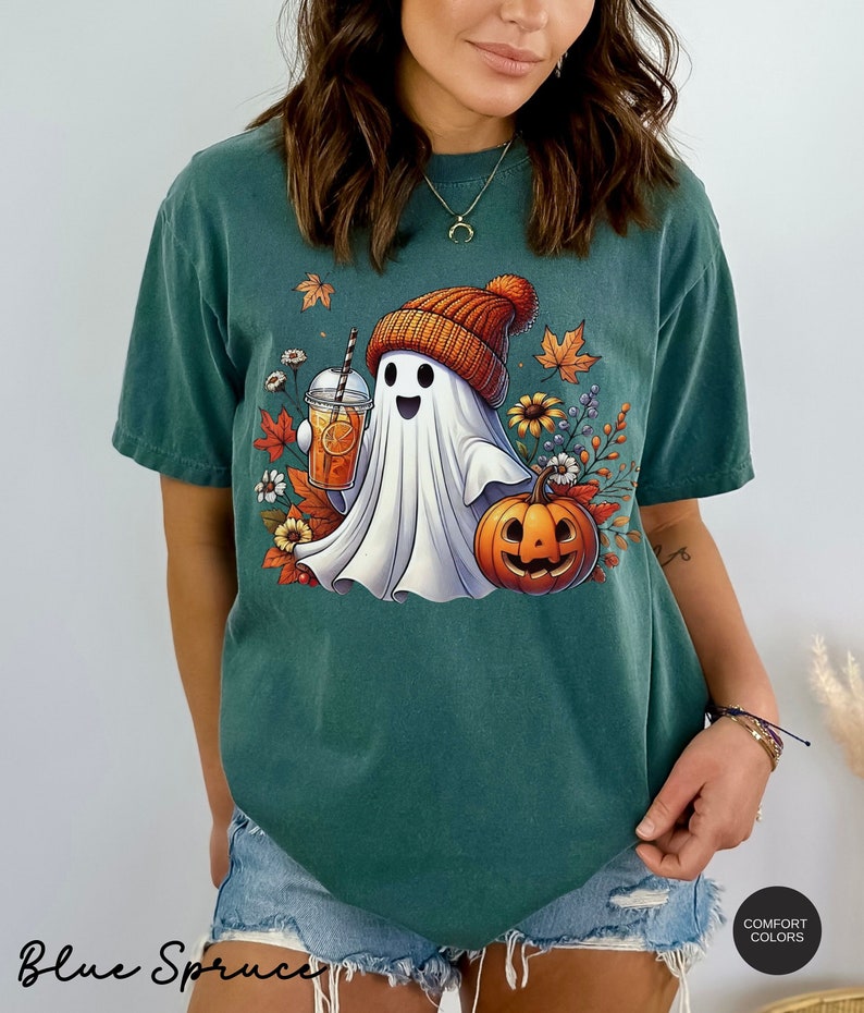 May include: A teal blue t-shirt with a white ghost wearing an orange and brown knit hat holding a glass of orange juice. The ghost is surrounded by fall leaves and flowers. The text "Blue Spruce" is visible at the bottom of the image.