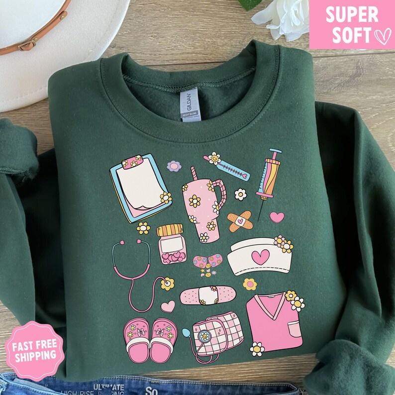 May include: A dark green sweatshirt featuring a collection of medical-themed illustrations in pink, yellow, and white. The design includes a stethoscope, syringe, prescription bottle, and a nurse's cap with a heart. The words "SUPER SOFT" are in a pink box.